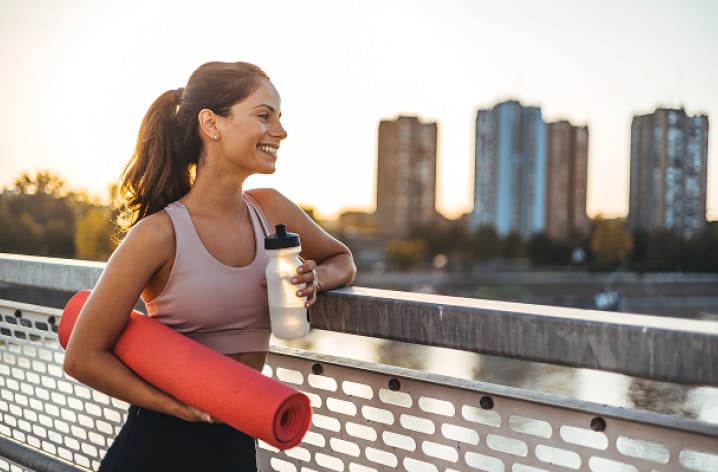 Frau in Sportkleidung mit Yogamatte und Trinkflasche lehnt an Brückengeländer, Skyline im Sonnenuntergang im Hintergrund