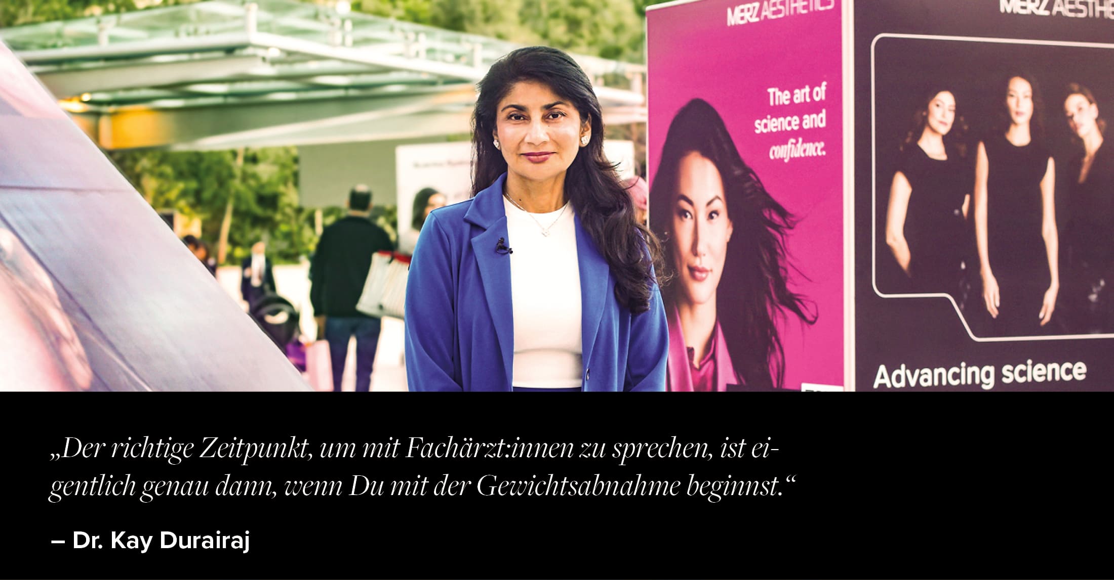 Dr. Kay Druairaj im blauen Blazer steht vor Kongressständen von Merz Aesthetics® mit Werbebannern, Kongressgelände im Hintergrund